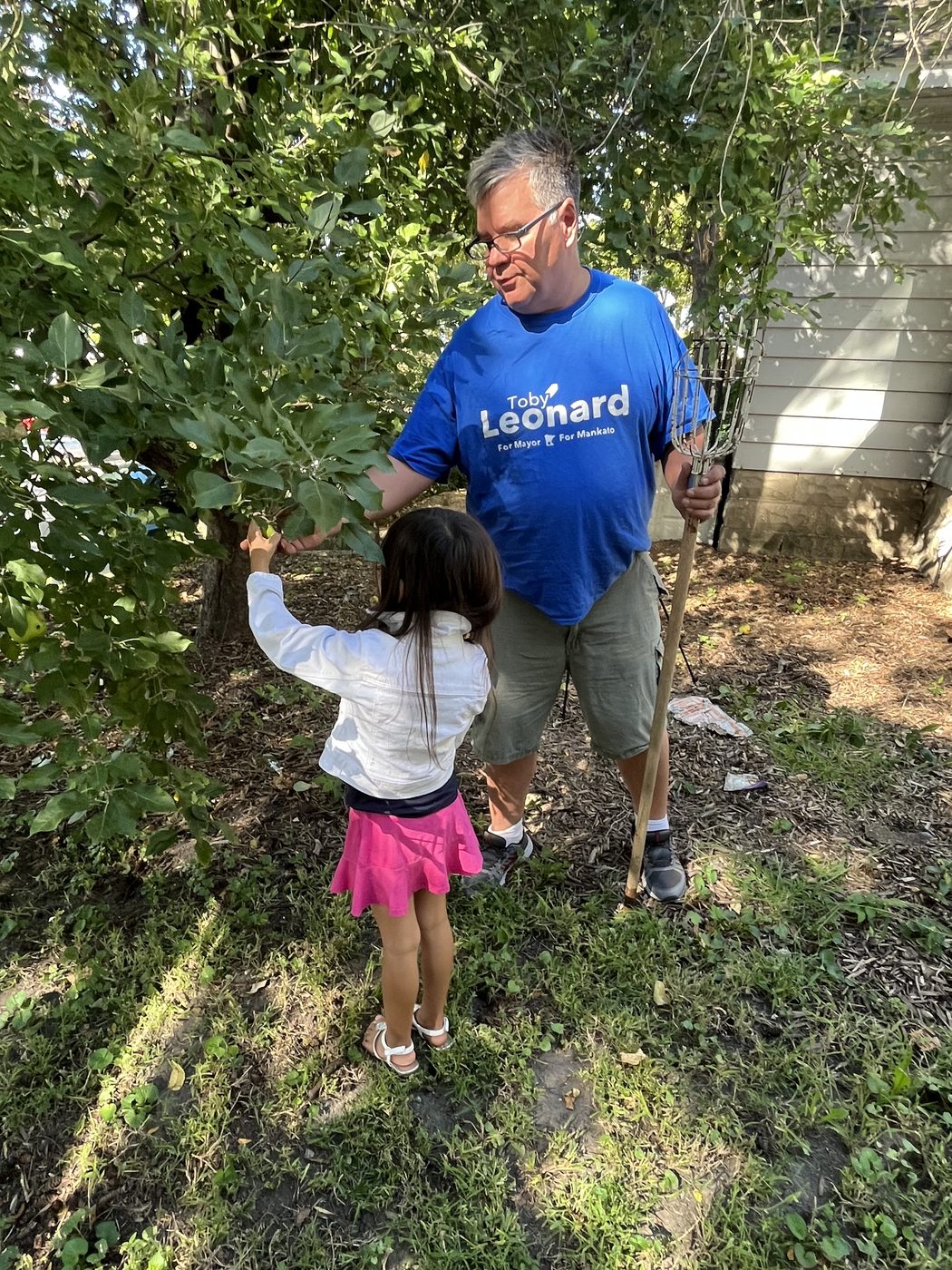 Toby Leonard in a campaign T-shirt holding a long-handled fruit picker while a young girl reaches for a branch