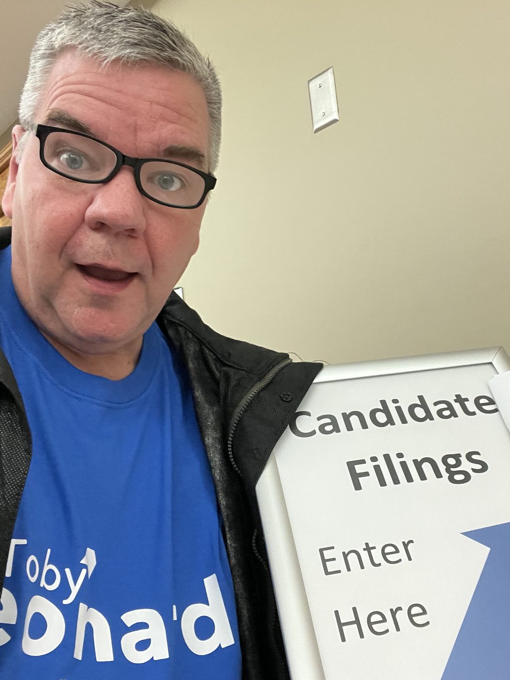 Toby Leonard in a campaign T-shirt taking a selfie beside a 'Candidate Filings — Enter Here' sign, campaign manual in hand