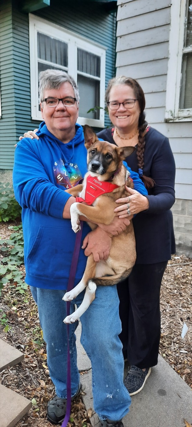 Toby Leonard holding a tan-and-white shepherd-mix dog in a red harness, outside a house